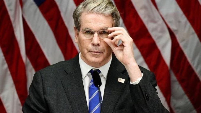 A man in a dark suit and blue tie, wearing a small American flag pin, adjusts his glasses while standing in front of U.S. flags. The image complements the post by visually representing Treasury leadership and the announcement of $36 billion in liquidity being injected into markets.