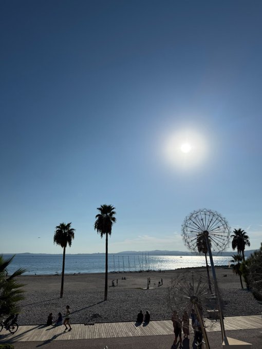 A beach scene at sunset featuring silhouettes of palm trees, a Ferris wheel, and people walking along the promenade with the sun reflecting on the water in the background.