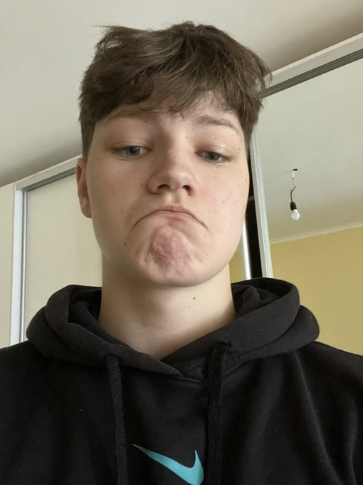 A young man wearing a black Nike hoodie makes a sad, pouty face at the camera in a casual, indoor setting with a mirror visible behind him.