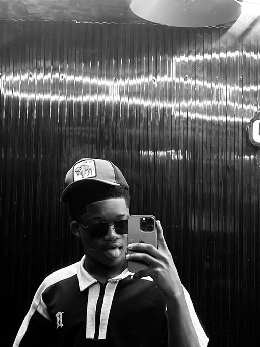 A black-and-white selfie of a young person wearing a baseball cap with a dog patch and sunglasses, holding up a smartphone to take the photo in front of a reflective, vertically-striped wall.