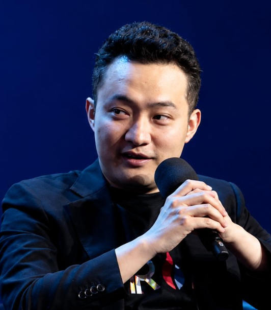A man in a black blazer speaks into a microphone at what appears to be a conference or panel event, set against a dark blue background. While the image itself shows a speaker mid-presentation, it seems unrelated to the post about Polygon's expansion of its Open Money Stack to integrate with TRON's stablecoin economy for payment services.
