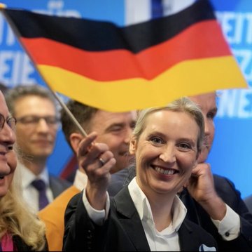 A smiling woman in a dark blazer holds up a German flag, surrounded by other people at what appears to be a political event.