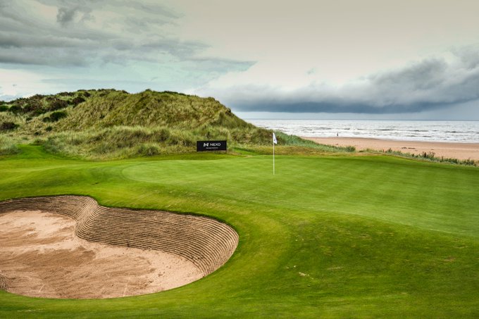 A scenic autumn view of a golf course green with a flagstick and bunker, surrounded by tall trees displaying vibrant orange and red foliage, with a sign reading "NEXO" visible among the trees, reflecting the Pro-Am event at Trump Scotland.