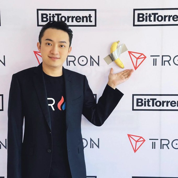 A man in a black blazer and TRON t-shirt holds a banana taped to a wall in front of a backdrop featuring BitTorrent and TRON logos.