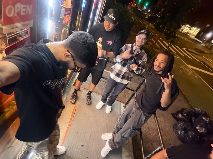 A nighttime street photo of four young men posing playfully outside a store with an "OPEN" sign, one wearing a LeBron Lakers cap and another making a hand gesture.