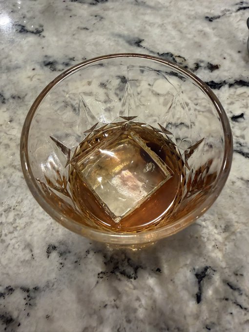 A rocks glass containing an amber-colored cocktail with a large square ice cube sits on a marble countertop.