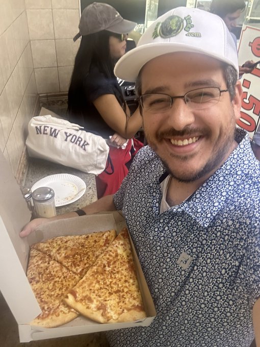 A man wearing glasses and a green "Boston" cap smiling while holding an open pizza box with slices inside, with a woman in a baseball cap visible in the background.