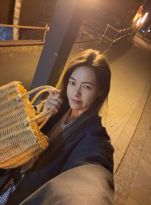 A nighttime selfie of a young woman with long dark hair holding a woven straw tote bag, with metal scaffolding visible in the background.
