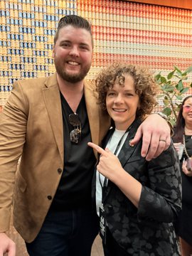 A bearded man in a tan blazer poses with his arm around a smiling woman with curly hair at what appears to be the Dog Talk event during DDNYC, with a colorful patterned backdrop behind them. The image visually represents the post's celebration of women leading the way in Web3, showcasing one of the "Dog Talk ladies" alongside a male collaborator in a moment of partnership and empowerment at the conference.