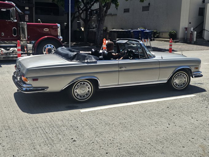 A silver classic Mercedes-Benz convertible drives down a city street with the top down, while a large red truck passes in the background.