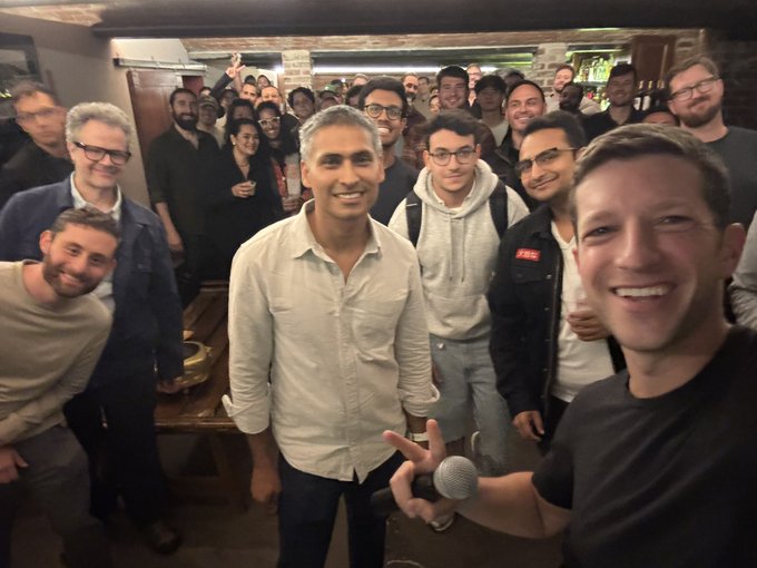 A man in the foreground takes a selfie holding a microphone while smiling broadly, with a large group of people gathered behind him in what appears to be a pub or event space, including a man in a white shirt at center, likely capturing the SF stablecoin community gathering referenced in the post.