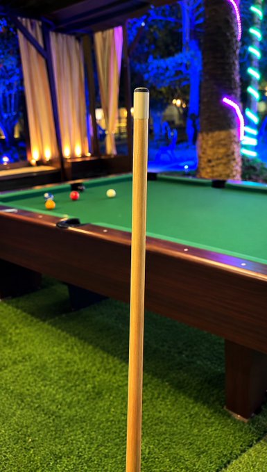 A pool cue stands upright in the foreground of an outdoor pool table with billiard balls scattered on the green felt, set against a backdrop of blue and purple lighting.
