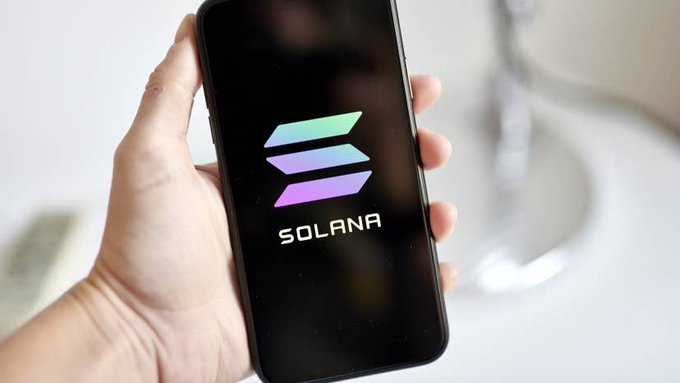A hand holding a smartphone displaying the Solana cryptocurrency logo on a black screen.