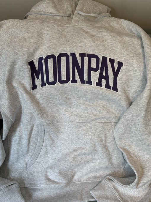 A heather gray pullover hoodie is laid out flat, featuring the word "MOONPAY" in large, arched purple varsity-style lettering across the chest, with a front kangaroo pocket visible below. The post's comment about it being a "nice hoodie" fits the image perfectly, as the item appears to be a casual, branded MoonPay merchandise piece.