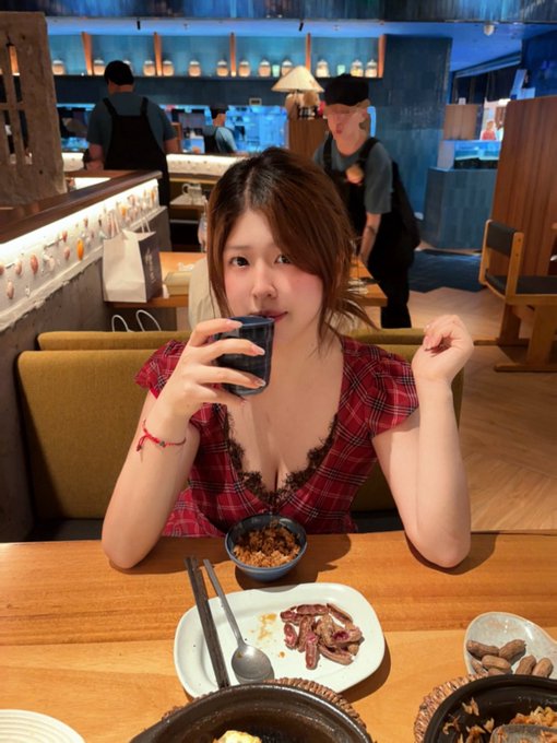 A young woman in a red plaid top drinks from a small dark cup at a restaurant table set with various dishes.