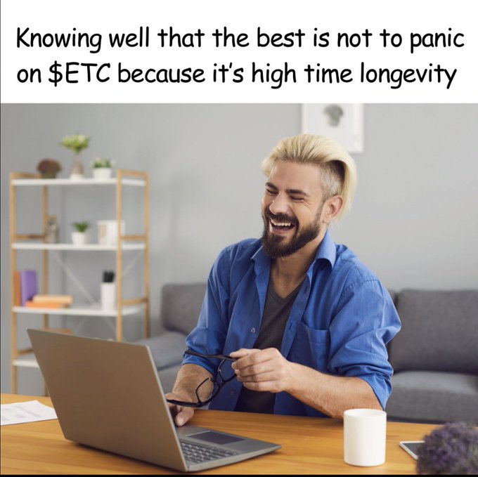 A blonde, bearded man in a blue shirt laughs while holding glasses at a laptop on a wooden desk in a bright living room. The text overlay reads, "Knowing well that the best is not to panic on $ETC because it's high time longevity," which visually reinforces the post's message about the long-term support and stability of the Ethereum Classic ($ETC) ecosystem.