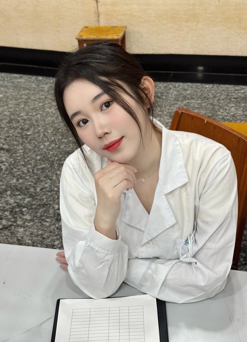 A young woman with long dark hair, wearing a white blouse and delicate necklace, rests her chin on her hand at a table with what appears to be a grid notebook. The post shares her journey from being a nurse to diving into Web3, and the image complements this narrative by showing her approachable, thoughtful demeanor as if she's ready to share her story and insights.