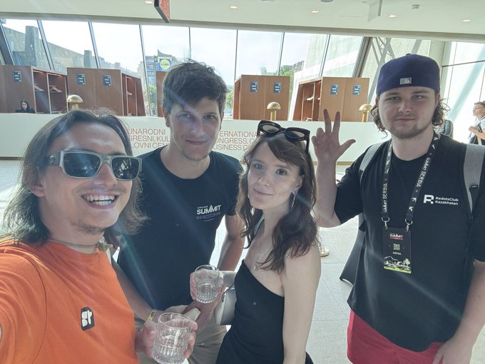 Four people pose for a photo at what appears to be a convention center, with a banner mentioning 'SUMMIT' visible behind them. One man wears a lanyard that reads 'RedwoodClub #askthebarn'.