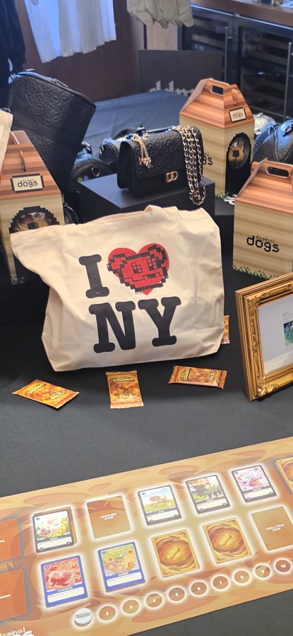 A retail display featuring an 'I ♥ NY' tote bag alongside designer handbags and trading card merchandise, with Pokémon-style cards laid out on a playmat below.