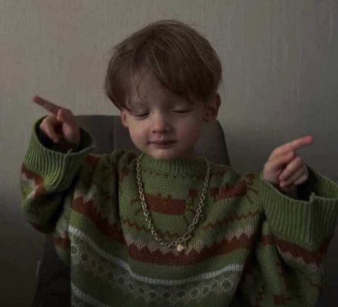 A young boy in a green and brown patterned sweater with a gold chain points both fingers while looking down.