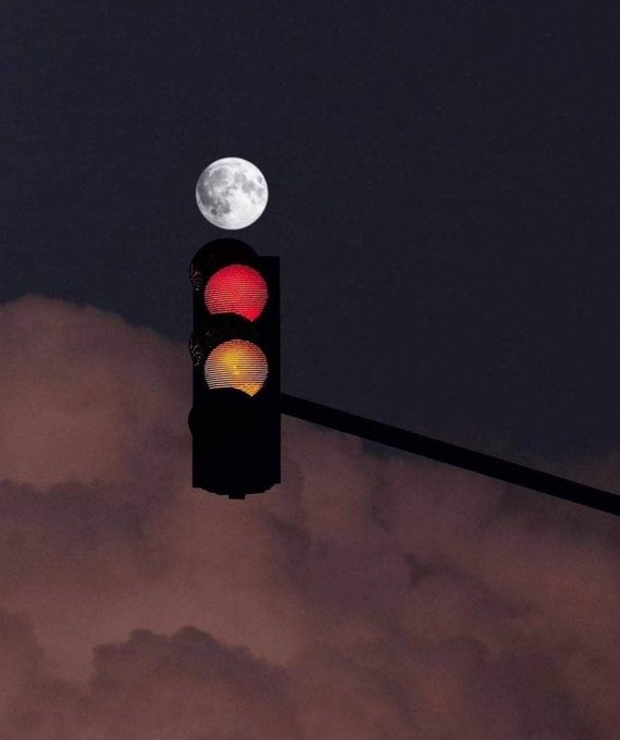 A glowing red traffic light hangs against a dark twilight sky, with a bright full moon positioned directly above the signal.