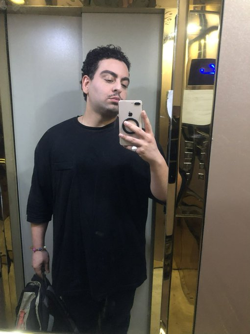A man with dark curly hair wearing a black t-shirt takes a mirror selfie with a white smartphone, holding a bag in his other hand.