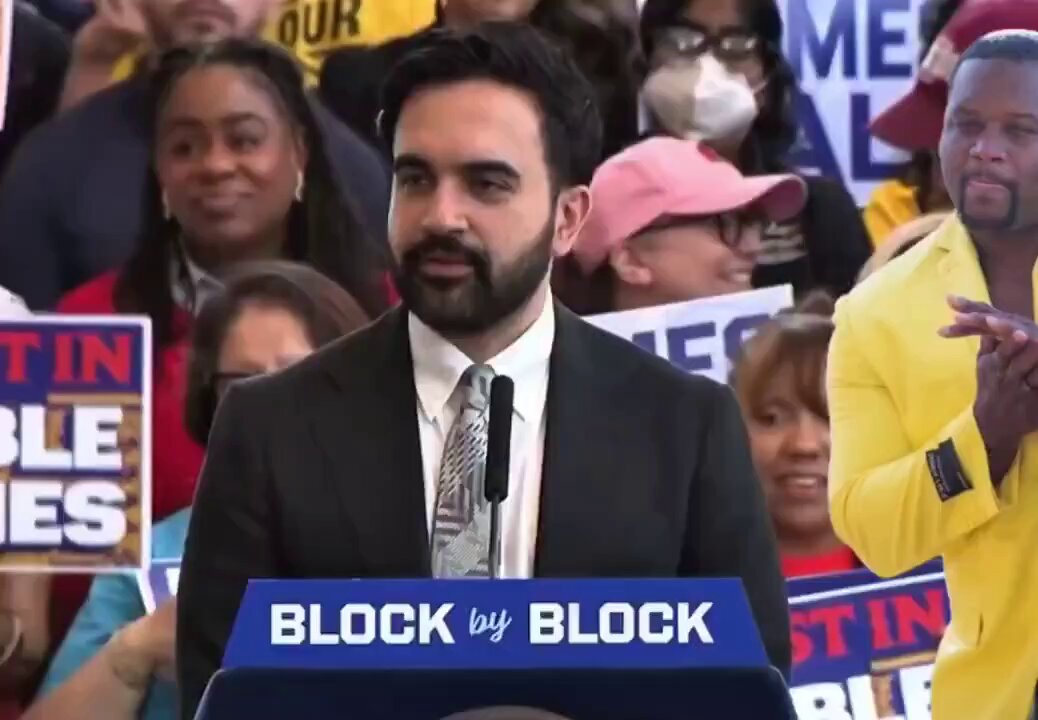 The image shows a man standing at a podium, giving a speech or presentation. He appears to be wearing a suit and tie, suggesting a formal event or gathering. The background of the image features a crowd of people, indicating that this could be an audience for his speech. There is also text in the image that reads "New Zcash ad just dropped", which might be related to the man's speech or the event taking place.