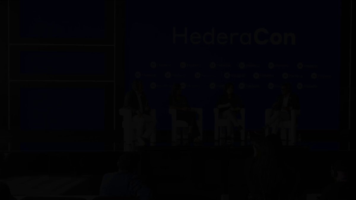 The image shows a group of people sitting around a table at an event, possibly a conference or panel discussion. The attendees are engaged in conversation, with some standing while others sit down. The setting appears to be indoors, as indicated by the presence of chairs and a stage. The background features a blue wall adorned with white text that reads "HederaCon 2026: agentic trust needs the proper foundation before it can scale. And it starts with verifiable data, authenticity, secure identity and open standards".