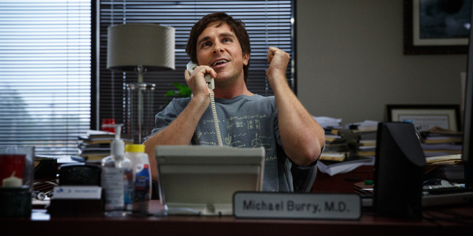 Christian Bale as Michael Burry in "The Big Short" sits at his desk with a phone, celebrating with a raised fist, with a nameplate reading "Michael Burry, M.D." visible. The image references the real-life investor behind the post's news about his new short positions in Nebius ($NBIS) and Oracle ($ORCL).
