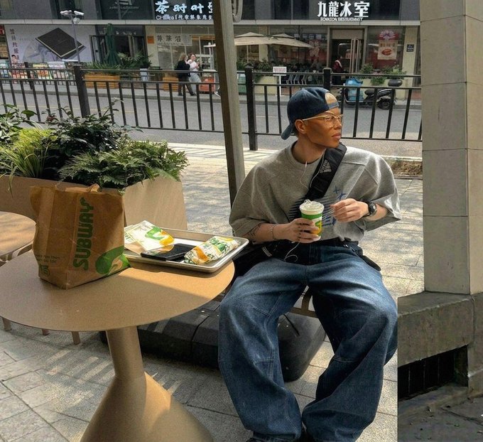 A stylish young man in a backwards cap, gray shirt, and baggy jeans sitting at an outdoor Subway table eating, with a Subway paper bag and food tray visible.