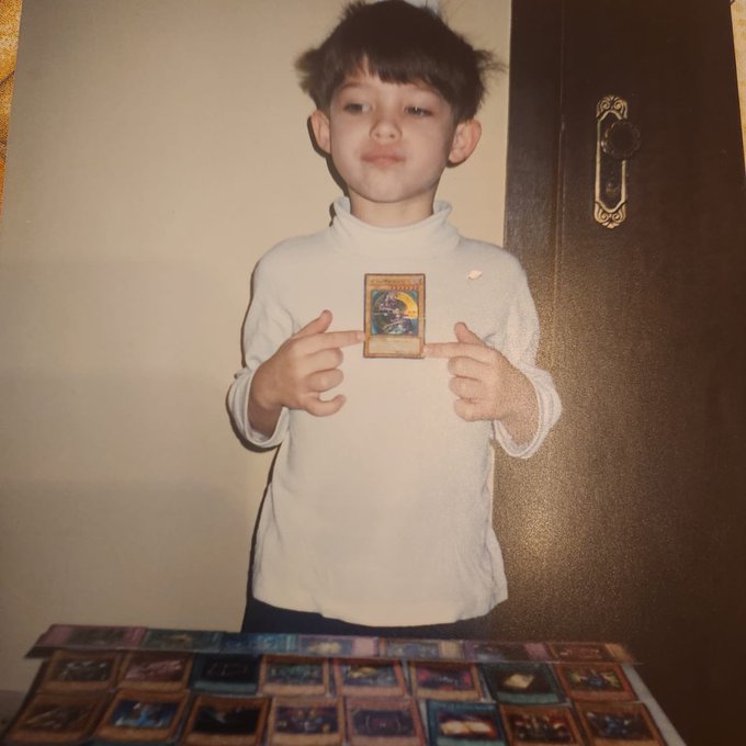 A vintage photograph of a young boy in a white turtleneck pointing at a trading card he's holding, with a spread of many trading cards laid out on a table in front of him.