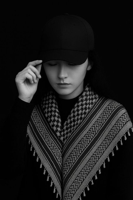 A black and white portrait of a young person wearing a baseball cap and a patterned keffiyeh-style scarf, eyes closed with their hand touching the brim of the cap.