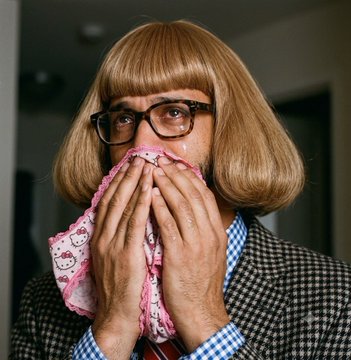A man wearing a brown bob wig, glasses, and a houndstooth jacket wiping his eyes with a pink handkerchief, appearing to cry.