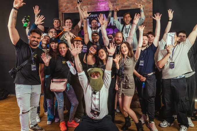 The image shows a group of people posing for a photo, with many of them raising their arms up in celebration or excitement. The group consists of at least 13 individuals standing close together against a black background. Some are wearing ties, indicating that they might be dressed formally for an event. The overall atmosphere appears to be joyful and energetic as the group poses for the picture.