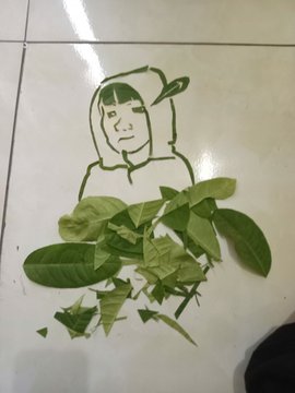A piece of green leaf art arranged on a white tiled floor to depict a simple human figure alongside scattered foliage.