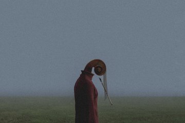A dark, atmospheric image of a figure in a red coat standing in a foggy field, holding a horned skull or mask to their face with horns hanging down.