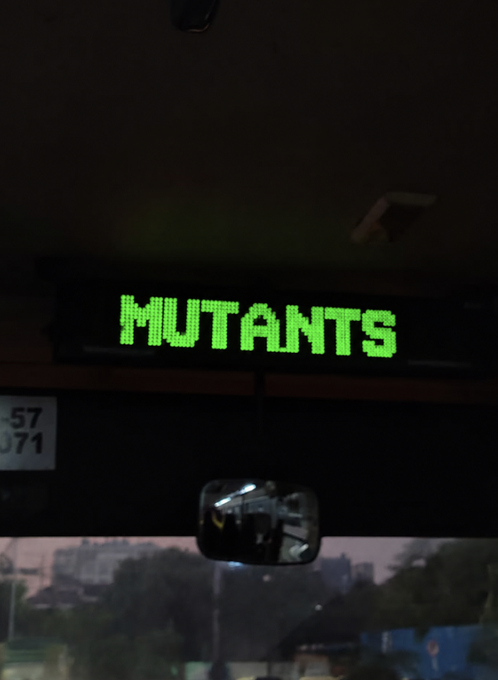 A green LED destination sign on a bus displays the word "MUTANTS," seemingly poking fun at fellow commuters, while the rearview mirror reflects a driver. The post's "HMM" reflects amusement or surprise at this unusual and eerie bus route designation.