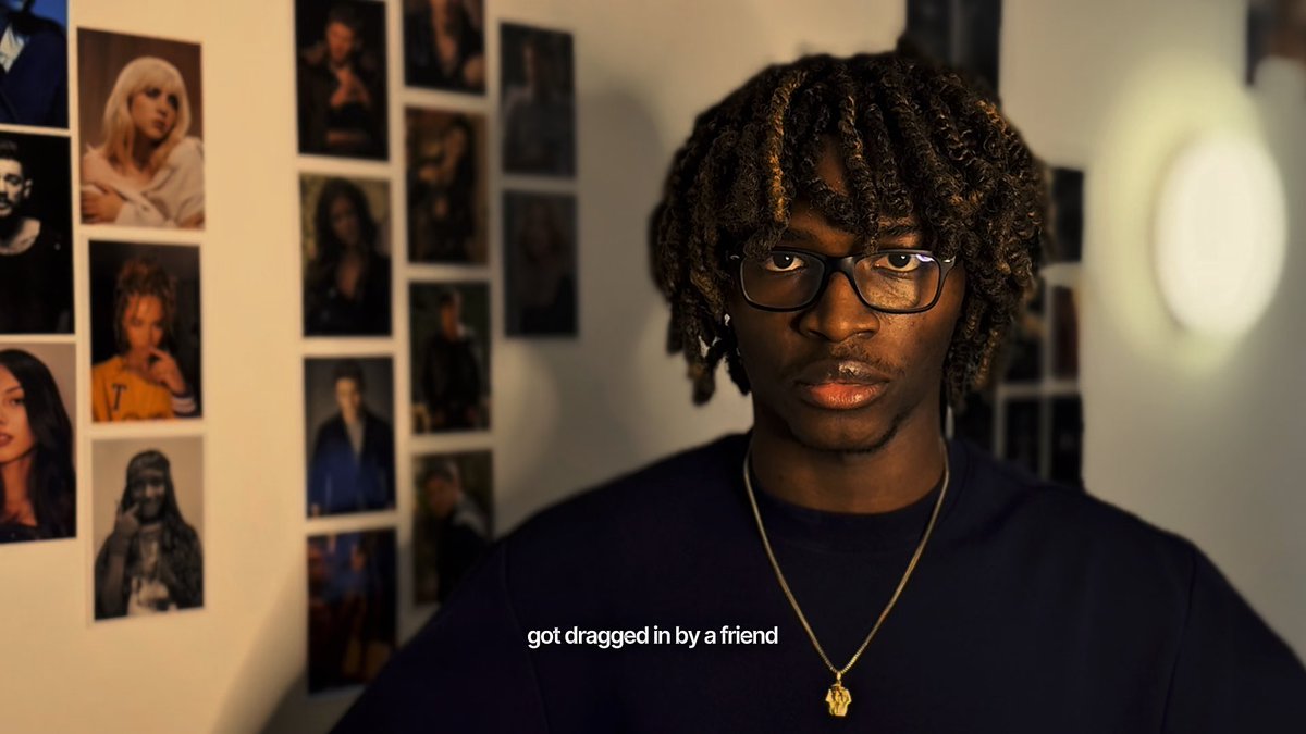 A young man with glasses and dreadlocks wearing a dark blue shirt and gold chain necklace stands in front of a wall covered with celebrity photos, with the subtitle 'got dragged in by a friend'.