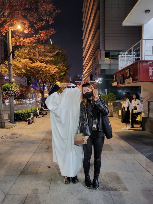 Two women pose on a city sidewalk at night; one wears a white ghost costume covering her face while the other wears a black leather jacket and face mask, making peace signs.