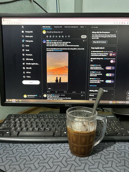 A glass mug of iced coffee with a spoon sits in front of a Dell monitor displaying the X (formerly Twitter) homepage in Vietnamese, with the "Dành cho bạn" (For You) feed visible. The image captures a morning productivity setup, perfectly matching the caption about enjoying coffee ("ly cf") to stay alert while scrolling through X.