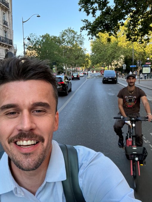 A smiling man in a light blue shirt takes a selfie on a city street, with another man riding a red e-bike visible behind him.