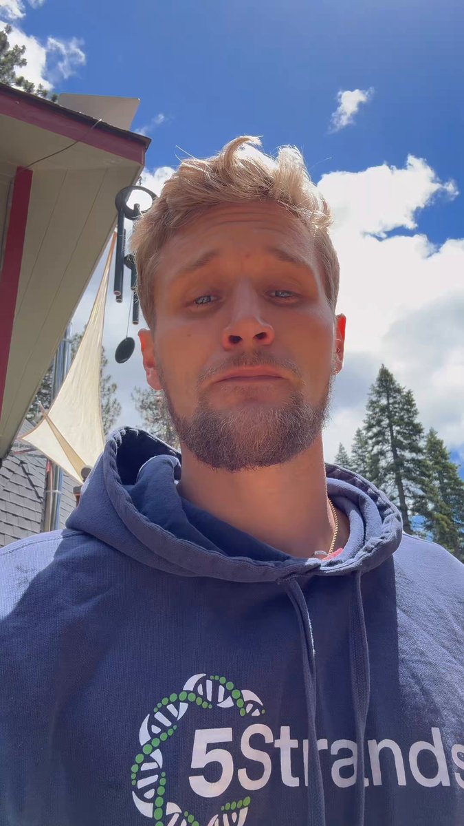 The image features a man wearing a blue hoodie with the words "5 Strands" printed on it, standing outside of a house under a cloudy sky. The man appears to be looking directly at the camera, possibly posing for a photo or video. There is also an element of text in the image that reads "New @solflare Merch Charity Event Merch @P4L_Puffsterz Secured a Few @solana Pieces Appreciate You @retrospect82". This suggests that the man might be involved in some sort of charitable event or promotion related to the 5 Strands brand.