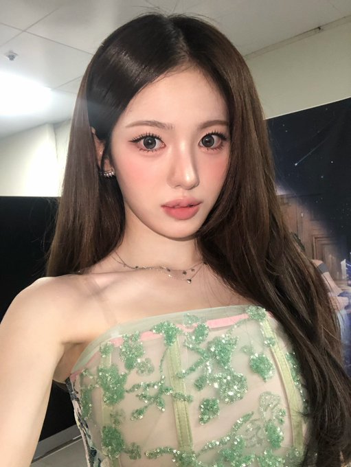 A selfie-style photo of a young woman with long brown hair wearing a sheer top with green floral embroidery, appearing to be backstage with stage lighting in the background.