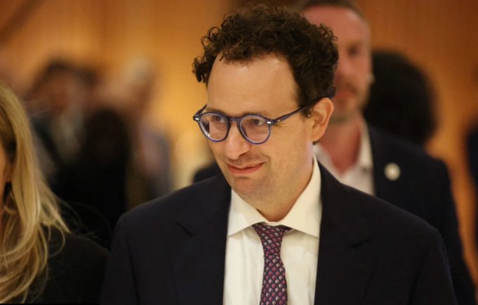 A man with curly hair and glasses, wearing a dark suit, white shirt, and patterned tie, smiles slightly while walking through a crowded indoor venue.