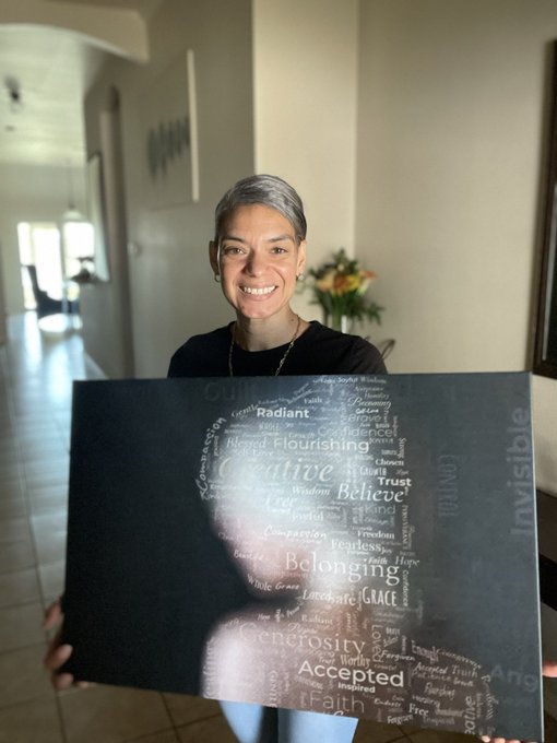 A smiling woman with short gray hair holds up a black canvas displaying a word art silhouette of a woman surrounded by words like 'Faith,' 'Creativity,' 'Grace,' and 'Belonging.'