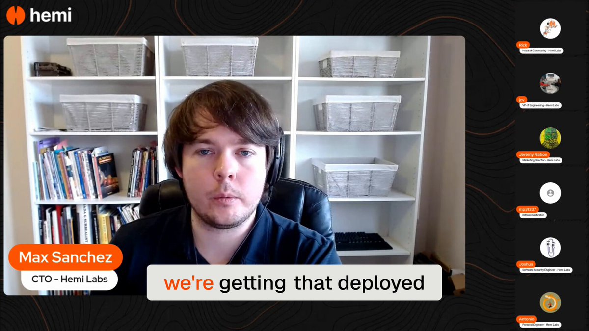 The image shows a man sitting at his desk in front of a computer screen, likely participating in an online meeting or webinar related to the deployment of the veHEMI contract upgrade. The man appears to be wearing headphones, suggesting that he is actively engaged in the session. There are several books scattered around him on the desk and shelves behind him, indicating that this might be his workspace or a shared office environment.