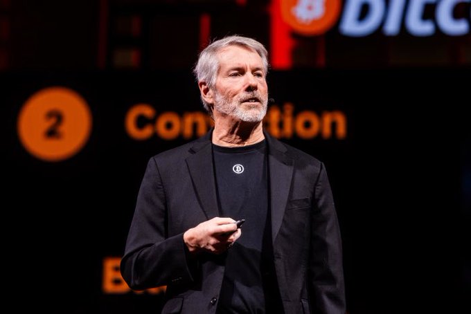 A man with gray hair and a beard speaking on stage at what appears to be a conference, holding a presentation clicker, with 'DIC' and '2 Convention' visible in the background.