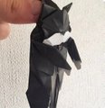 A hand holds a small black origami figure against a white wall.