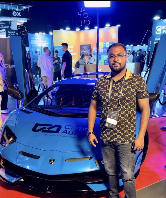 A man wearing glasses and a patterned shirt stands in front of a blue Lamborghini at a CryptoAutos exhibition, with the company's website visible on the car.
