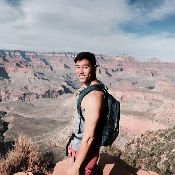 A smiling young man wearing a gray tank top and backpack poses at the edge of a canyon, likely the Grand Canyon, with expansive rocky landscapes behind him.